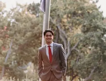 Man with black hair and tan suit standing in front of flagpole